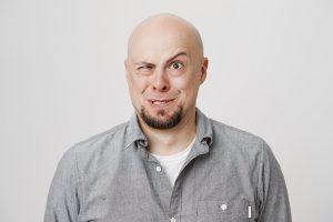 Indoor portrait of funny bald european man making crazy face while standing over gray background. Actor performes on stage in play for children. Guy acts weird to avoid conversation with police